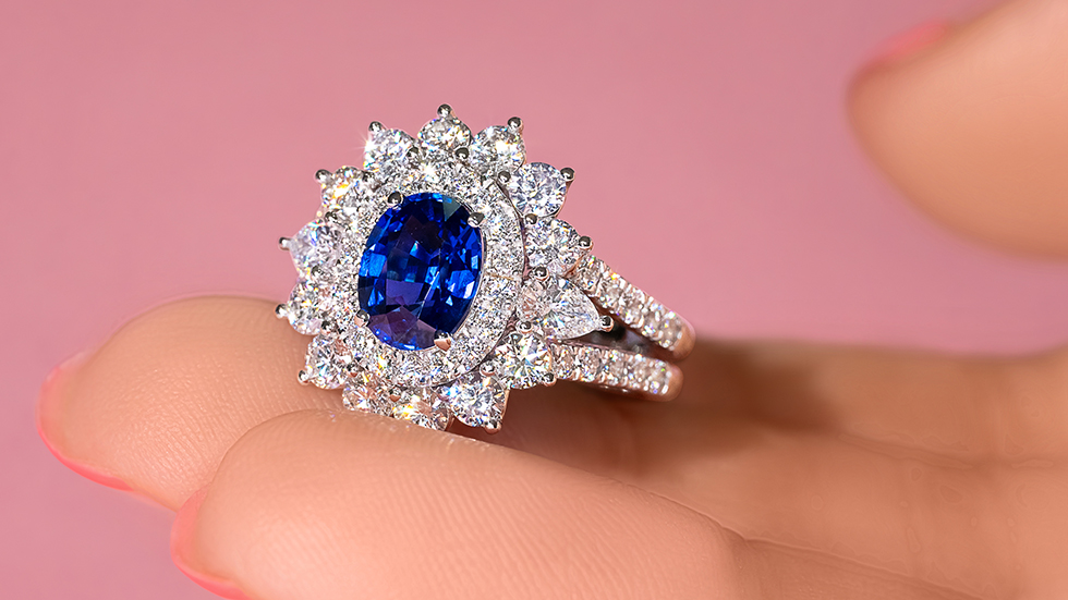 A photo of a manicured hand holding up an unheated blue Sapphire ring, encircled with two diamond halos, and a double band.